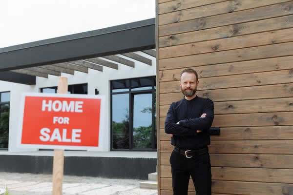 man dressed in black standing beside a house for sale sign
